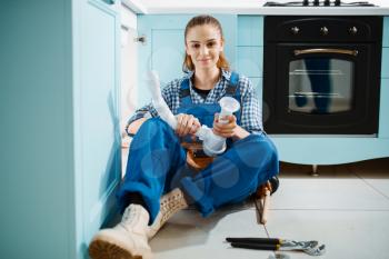 Cute female plumber in uniform holds drain pipe in the kitchen. Handywoman with toolbag repair sink, sanitary equipment service at home