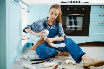 Attractive female plumber in uniform fixing problem with drain pipe in the kitchen. Handywoman with toolbag repair sink, sanitary equipment service at home