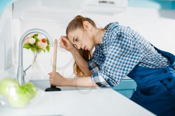 Tired female plumber in uniform holds plunger, clog in the kitchen. Handywoman repair sink, sanitary equipment service at home