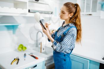 Cute female furniture maker in uniform holds notebook in the kitchen. Handywoman fixing problem with garniture cabinet, repairing service at home