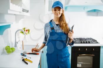 Cute female furniture maker in uniform holds notebook in the kitchen. Handywoman fixing problem with garniture cabinet, repairing service at home