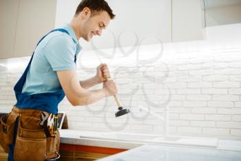 Male plumber in uniform holds plunger, problem with clog in the kitchen. Handyman with toolbag repair sink, sanitary equipment service at home