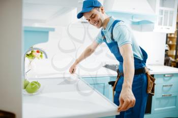Male furniture maker in uniform measures table top in the kitchen. Handyman installing garniture, repairing service at home