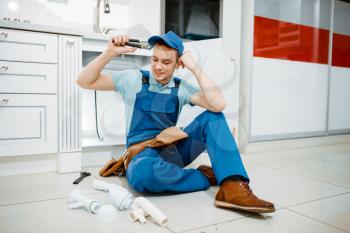 Smiling male plumber in uniform holds drain pipe in the kitchen. Handywoman with toolbag repair sink, sanitary equipment service at home