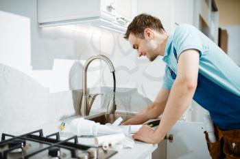 Male plumber in uniform fixing problem with faucet in the kitchen. Handyman with toolbag repair sink, sanitary equipment service at home