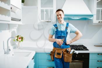 Cheerful male plumber in uniform poses in the kitchen. Handyman with toolbag repair sink, sanitary equipment service at home