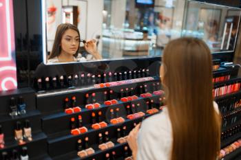 Woman applies mascara at the mirror in cosmetics store. Buyer at the showcase in luxury beauty shop salon, female customer in fashion market