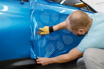 Male mechanic installs protective vinyl foil or film on vehicle door. Worker makes auto detailing. Automobile paint protection, professional tuning
