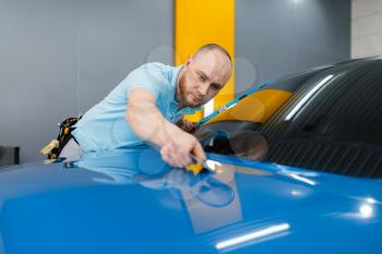 Male car wrapper with squeegee installs protective vinyl foil or film on hood. Worker makes auto detailing. Automobile paint protection coating, professional tuning