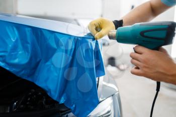 Car wrapping, man with drier installs protective vinyl foil or film on vehicle hood closeup. Worker makes auto detailing. Automobile paint protection coating, professional tuning