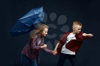 Little boy and girl with umbrella against powerful airflow in studio, wind effect, windy. Children with developing hairs, kids isolated on dark background, child emotion