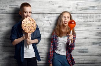 Little boy and girl holds big lollipops in studio. Children and sweets, kids isolated on wooden background, child photo session