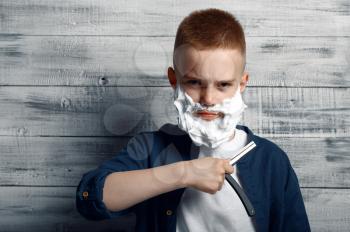 Serious little boy with shaving foam on his face holds a razor in studio. Kid isolated on wooden background, child emotion, schoolboy photo session
