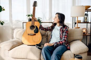 Cute woman with guitar at home, headphones on background. Pretty lady with musical instrument relax in the room, female music lover resting