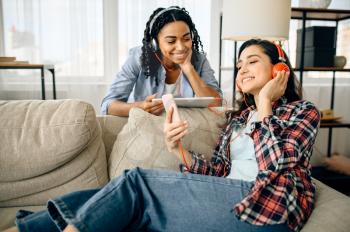 Two happy women enjoys listening to music indoors. Pretty girlfriends in earphones relax in the room, sound lovers resting on couch, female friends leisures together