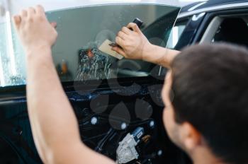 Male worker installs wetted car tinting, tuning service. Mechanic applying vinyl tint on vehicle window in garage, tinted glass