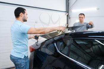 Male workers holds sheet of car tinting, tuning service. Mechanics applying vinyl tint on vehicle window in garage, tinted automobile glass