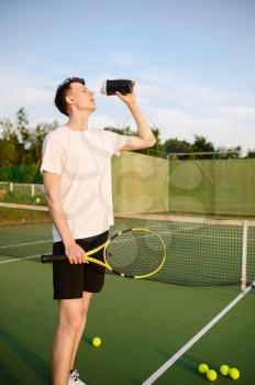 Tired male tennis player drinks water on outdoor court. Active healthy lifestyle, sport game competition, fitness training with racquet