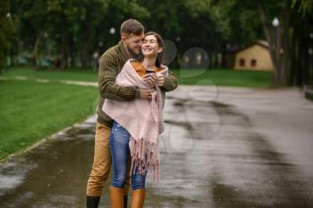 Love couple bask against each other in park, summer rainy day. Man and woman stand under umbrella in rain, romantic date on walking path, wet weather in alley
