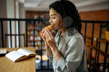 Cute female student eats croissants in cafe. Woman learning a subject in coffeehouse, education and food. Girl studying in campus cafeteria