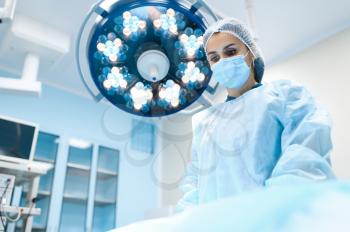 Female surgeon in gown and mask in operating room, lamp on background, surgery operation. Doctor in uniform, medical clinic worker, medicine and health, healthcare in hospital