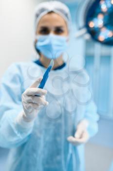 Female surgeon in gown and mask shows scalpel in operating room, lamp on background, surgery operation. Doctor in uniform, medical clinic worker, surgical medicine and health, healthcare in hospital