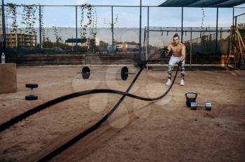 Strong man doing exercise with ropes, street workout, crossfit. Fitness training on sports ground outdoor, male person pumps muscles