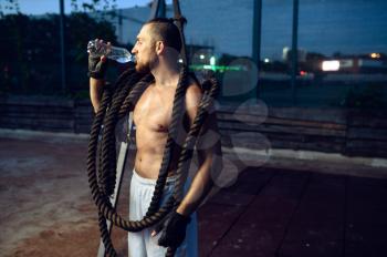 Muscular man with crossfit ropes drinks water, street workout. Fitness training on sports ground outdoor, male person pumps muscles, active urban lifestyle