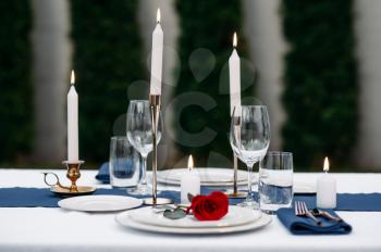 Table setting, wineglasses, candles and flower on the plate closeup, nobody. Luxury silverware and white tablecloth, tableware outdoors