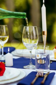 Table setting, wine is poured from a bottle into a wineglass, candles and flower on the plate closeup, nobody. Luxury silverware and white tablecloth, tableware outdoors