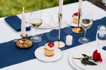 Table setting, glasses, candles, flower and sweets on the plates closeup, nobody. Luxury silverware and white tablecloth, tableware outdoors