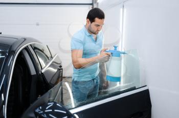 Male wrapper with spray prepares car for tinting, tuning service. Worker applying vinyl tint on vehicle window in garage, tinted automobile glass