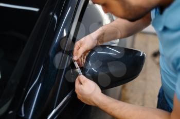 Male worker cuts film, car tinting installation process, tuning service. Worker applying vinyl tint on vehicle window in garage, tinted automobile glass