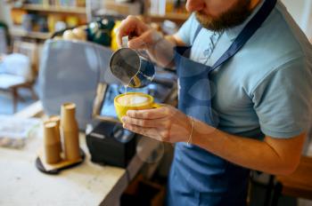 Male barista in apron pours foam into the coffee in cafe. Man makes fresh espresso in cafeteria, waiter at the counter in bar