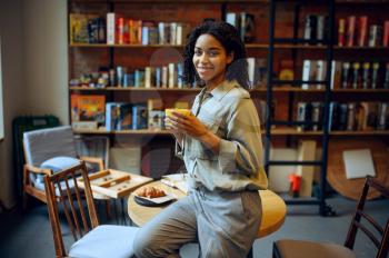 Smiling female student in library cafe. Woman with coffee and croissants poses at the table , education and knowledge. Girl studying in campus