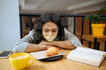 Pretty female student with her mouth taped shut looking on croissant in library cafe. Woman learning a subject, education and knowledge. Girl studying in campus cafeteria