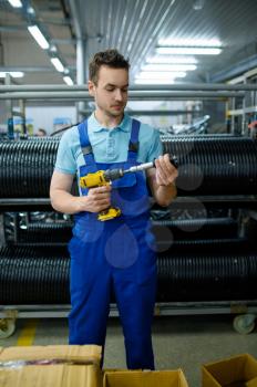 Worker in uniform holds electric screwdriver and bicycle hub, rows of tires on background. Bike wheels assembly on factory, cycle parts installation