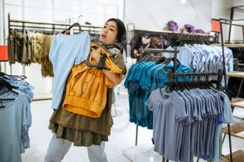 Young customer holds clothes on hanger in clothing store. Female person shopping in fashion boutique, shopaholic, shopper looking on garment