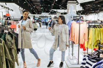 Two girls trying on coats in clothing store. Women shopping in fashion boutique, shopaholics, shoppers looking garment on hangers
