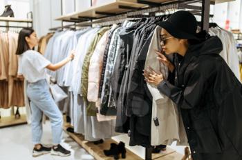 Two cute girls buying clothes in clothing store. Women shopping in fashion boutique, shopaholics, shoppers looking garment on hangers