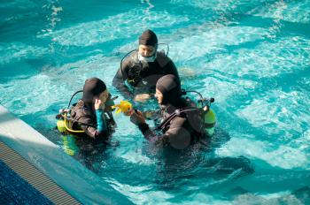 Instructor and two divers in aqualungs, dive course in diving school. Teaching people to swim underwater with scuba gear, indoor swimming pool interior on background, group training