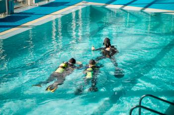 Divemaster and two divers in aqualungs, dive course in diving school. Teaching people to swim underwater with scuba gear, indoor swimming pool interior on background, group training