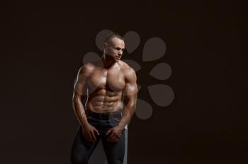 Male muscular athlete sitting on cube in studio, dark background. One man with athletic build, shirtless sportsman in jeans pants, active healthy lifestyle
