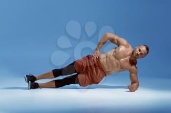 Male athlete keeps his balance on his elbow, workout in studio, blue background. One man with athletic build, shirtless sportsman in sportswear, active healthy lifestyle