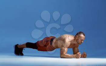 Male athlete trains his abs standing on his elbows, workout in studio, blue background. One man with athletic build, shirtless sportsman in sportswear, active healthy lifestyle