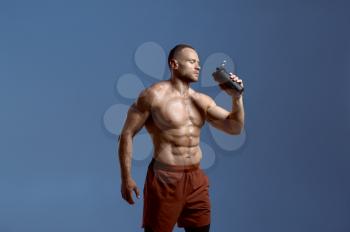 Muscular male athlete drinks water from sports bottle, photo shoot in studio, blue background. One man with athletic build, shirtless sportsman in sportswear, active healthy lifestyle