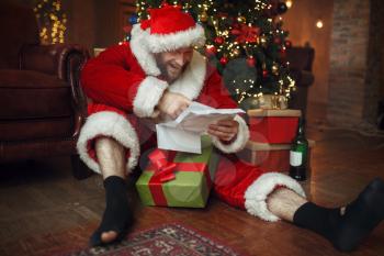 Bad drunk Santa claus reads letters under christmas tree, nasty party, humor. Unhealthy lifestyle, bearded man in holiday costume, new year and alcoholism