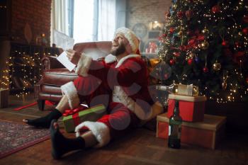 Bad drunk Santa claus reads letters under christmas tree, nasty party, humor. Unhealthy lifestyle, bearded man in holiday costume, new year