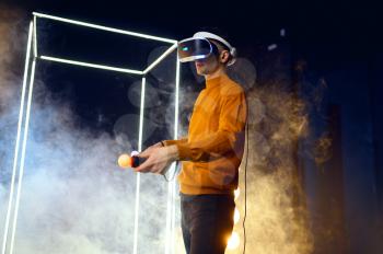 Male gamer plays the game using virtual reality headset and gamepad in luminous cube. Dark playing club interior, spotlight on background, VR technology with 3D vision