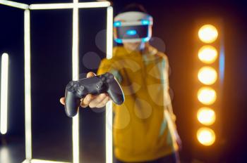 Man leisures using virtual reality headset and gamepad in luminous cube, front view. Dark playing club interior, spotlight on background, VR technology with 3D vision
